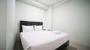 Un lit bien fait, avec du linge de lit blanc et deux serviettes enroulées, une tête de lit sombre et une petite table de chevet.