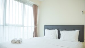 A neatly made bed with white linens and two rolled towels, a dark headboard, and a window with sheer curtains.