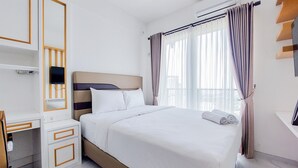 A hotel room with a bed, a desk with drawers, a television mounted on the wall, and a window with curtains. - Relaxing Studio At Sky House Bsd Apartment (Pagedangan)