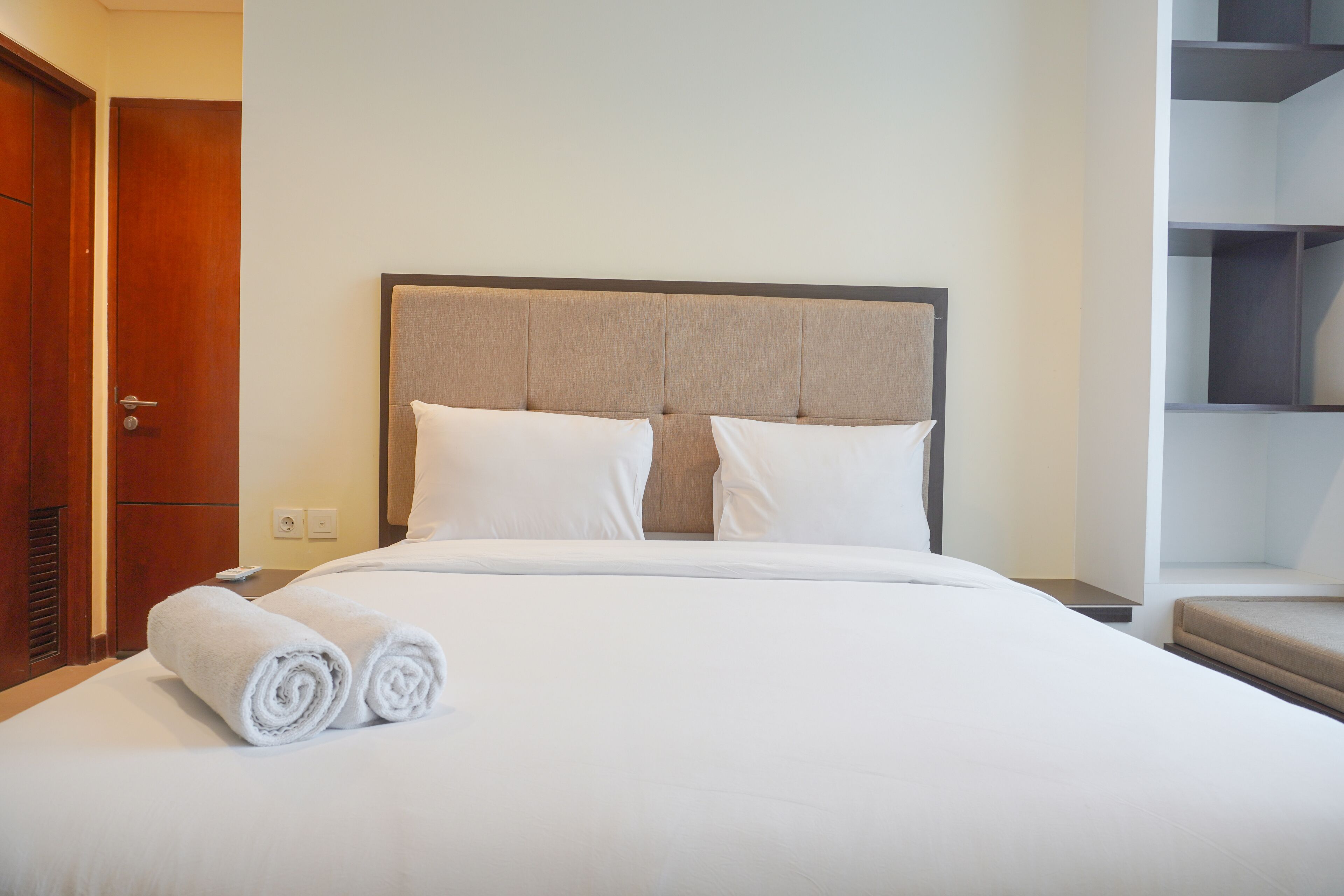 A neatly made bed with white linens and two rolled towels, a wooden door, and a built-in shelving unit to the right.