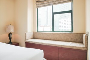 Habitación de hotel con cama, mesita de noche, lámpara, ventana con vista a edificios y un banco junto a la ventana.