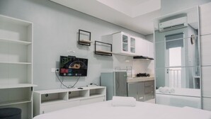 Moderni hotellihuone, jossa on sänky, televisio, pieni keittonurkkaus ja lasiseinällä erotettu kylpyhuone.