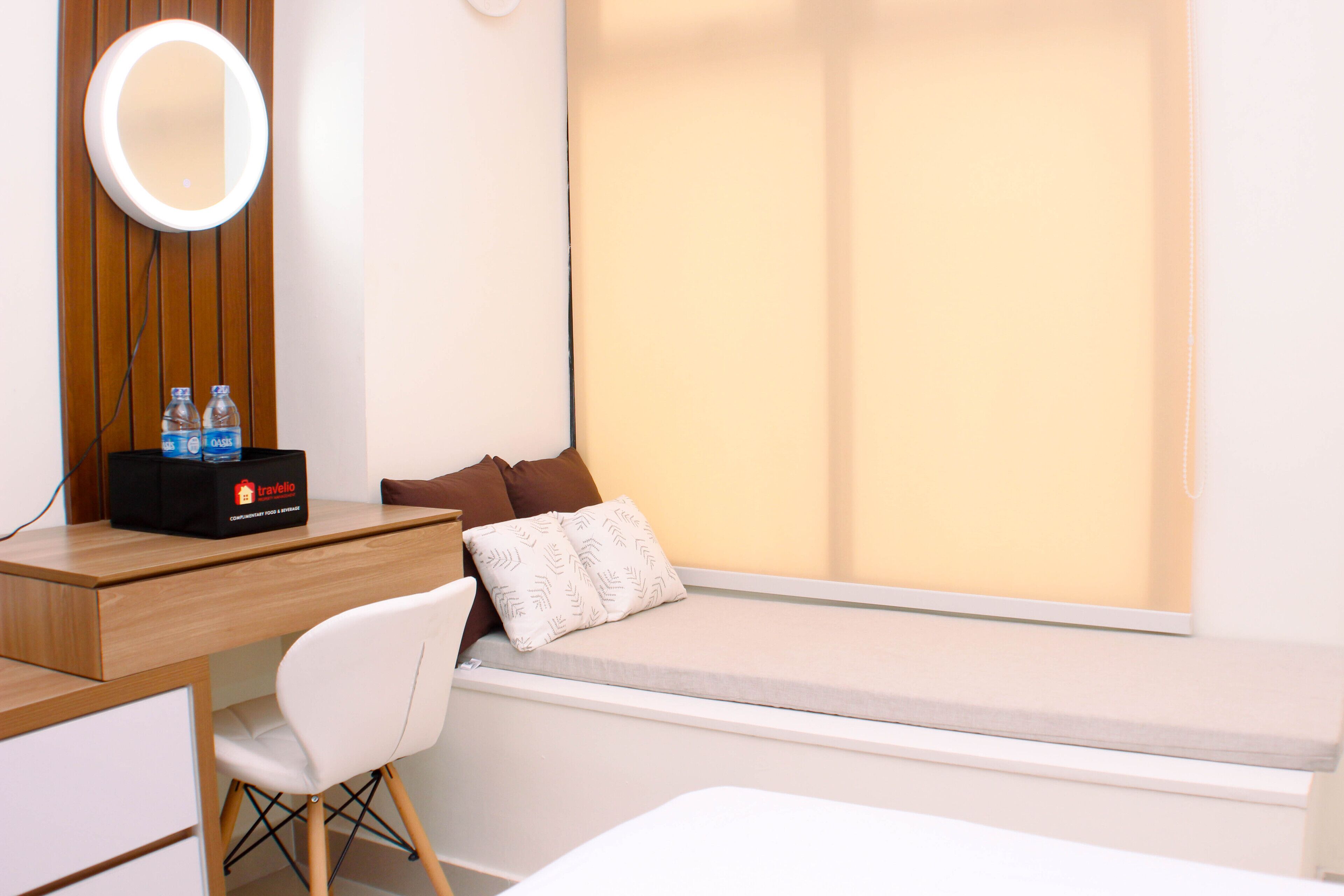 A compact hotel room with a wooden desk, a white chair, a small bed, a bedside table with a mirror, and a window with blinds.