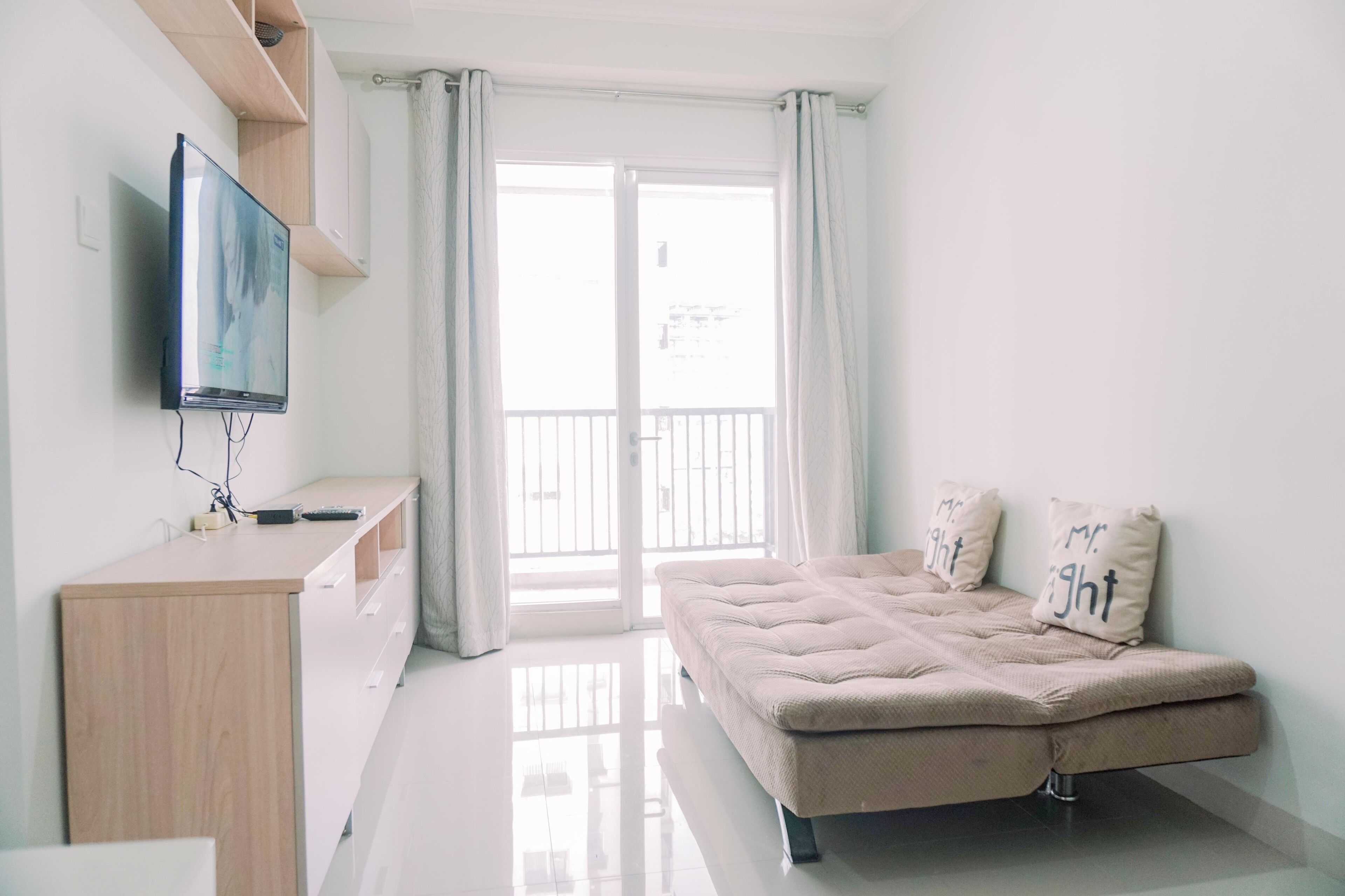 A modern bedroom with a sofa bed, a TV mounted on the wall, and a sliding glass door leading to a balcony.