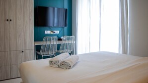 A hotel room with a bed, a TV mounted on the wall, a desk with two chairs, and a window with curtains. - Well Appointed And Homey Studio At West Vista Apartment (Jakarta)