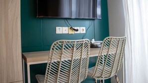 A room with a teal wall, a flat-screen TV mounted above a wooden desk, two wicker chairs, and a power outlet with a plug. - Well Appointed And Homey Studio At West Vista Apartment (Jakarta)