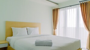A neatly made bed with white linens and a folded towel, a wooden headboard, and a nightstand with a lamp. - Comfortable And Minimalist Studio At Amethyst Apartment (Jakarta)