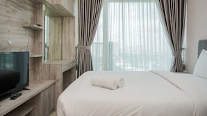 A hotel room with a wooden wall, a TV on a wooden shelf, a bed with white linens, and a window with curtains. - Minimalist And Nice Studio At Tree Park City Cikokol Apartment (Tangerang)