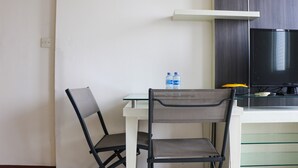 Quarto pequeno e moderno com escrivaninha branca, duas cadeiras pretas, mesa de vidro, garrafa de água, televisão e armário de madeira.