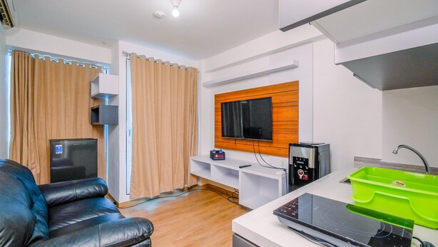 A modern living room with a black leather sofa, a flat-screen TV mounted on a wooden panel, a built-in sound system, and a kitchen area with a green sink and a stove. - Comfortable And Warm 2Br At Tifolia Apartment (Jakarta)
