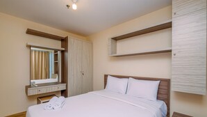 A neatly made bed with white linens, a wooden headboard, and a bedside table with a drawer. A mirror hangs above the bed, and a wooden shelf is mounted on the wall. - Comfortable And Warm 2Br At Tifolia Apartment (Jakarta)