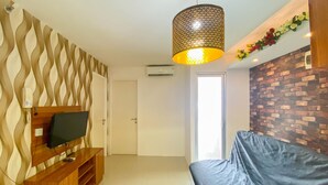 A living room with a TV on a wooden stand, a black sofa, a patterned wall, a brick wall, and a hanging light fixture. - Comfort And Homey 3Br Bassura City Apartment (Jakarta)