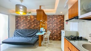 A compact living space with a sofa, a small dining table with chairs, a kitchen area with a stove, and a hanging light fixture. - Comfort And Homey 3Br Bassura City Apartment (Jakarta)