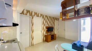 Habitación de hotel compacta con kitchenette, comedor con mesa de vidrio, televisor montado en una pared decorada y ventana que proporciona luz natural.