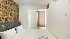 A hotel room with a bed, white bedding, a headboard, a window with curtains, and a wall with a patterned design. - Comfort And Homey 3Br Bassura City Apartment (Jakarta)