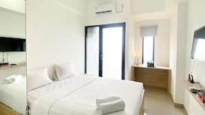 A hotel room with a large bed, a bedside table with a lamp, a TV, and an air conditioning unit. - Comfort And Cozy Designed Studio Sayana Bekasi Apartment (Tarumajaya)