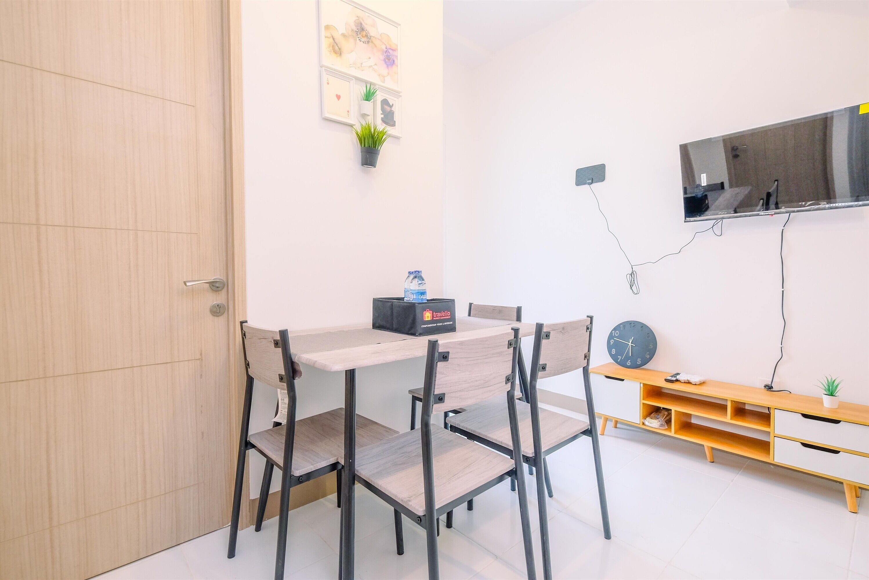 Um quarto com porta de madeira, uma mesa com cadeiras, uma televisão fixada na parede e uma pequena estante com uma planta.
