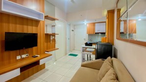 A modern living room with a flat-screen TV, wooden paneling, a beige sofa, and a kitchen area in the background. - Restful And Tidy 2Br Apartment Springlake Summarecon Bekasi (Bekasi)