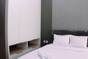 Un lit bien fait, avec du linge de lit blanc et des oreillers, une table de chevet en bois et une armoire intégrée dotée d’étagères ouvertes.