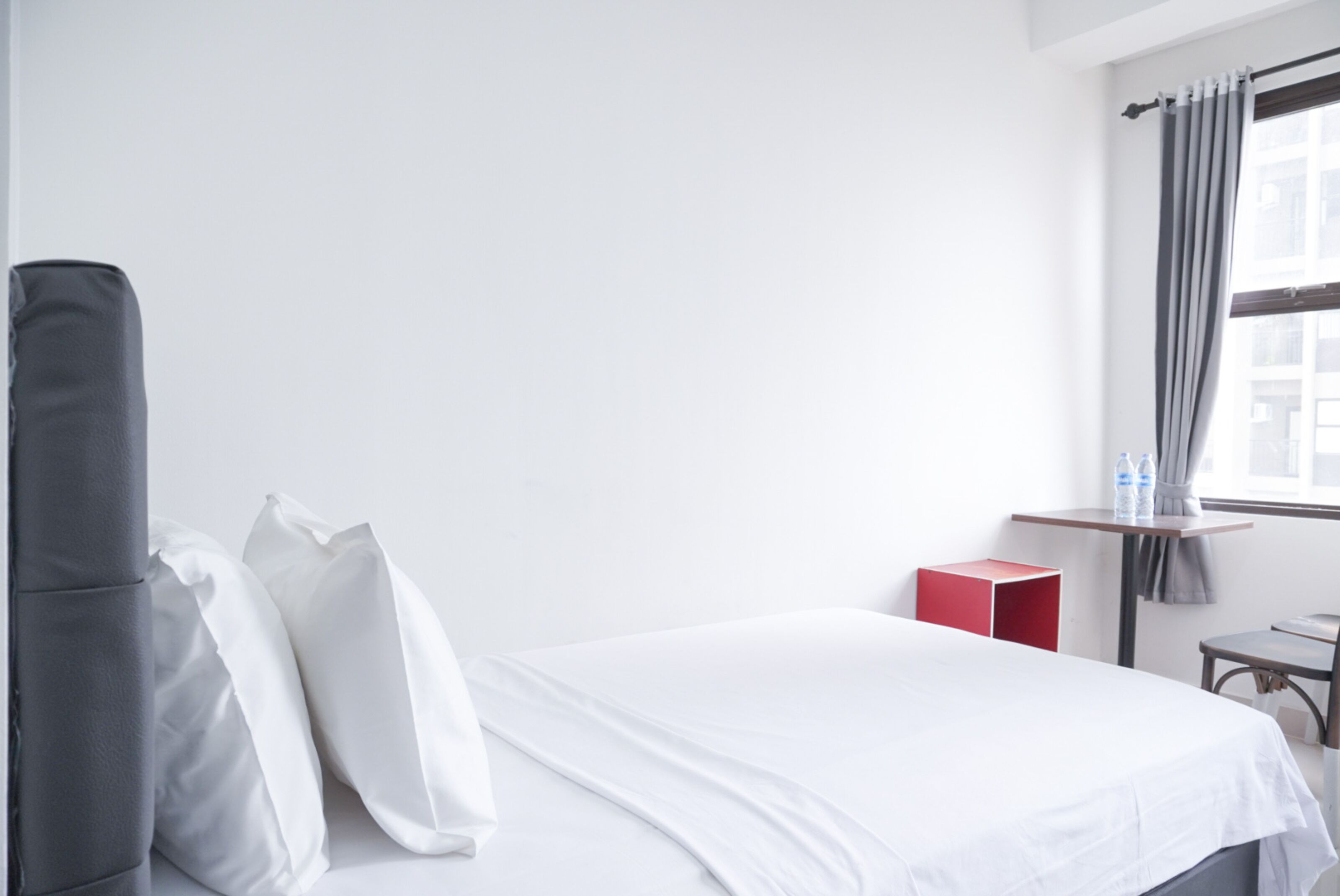 Ein Hotelzimmer mit Bett, Kissen, einer roten Kiste, einem kleinen Tisch und einem Fenster mit Vorhängen.