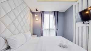 A hotel room with a tufted headboard, a flat-screen TV mounted on the wall, a neatly made bed with white linens, and a window with sheer curtains. - Comfortable And Minimalist 1Br At The Ayoma Apartment (South Tangerang)