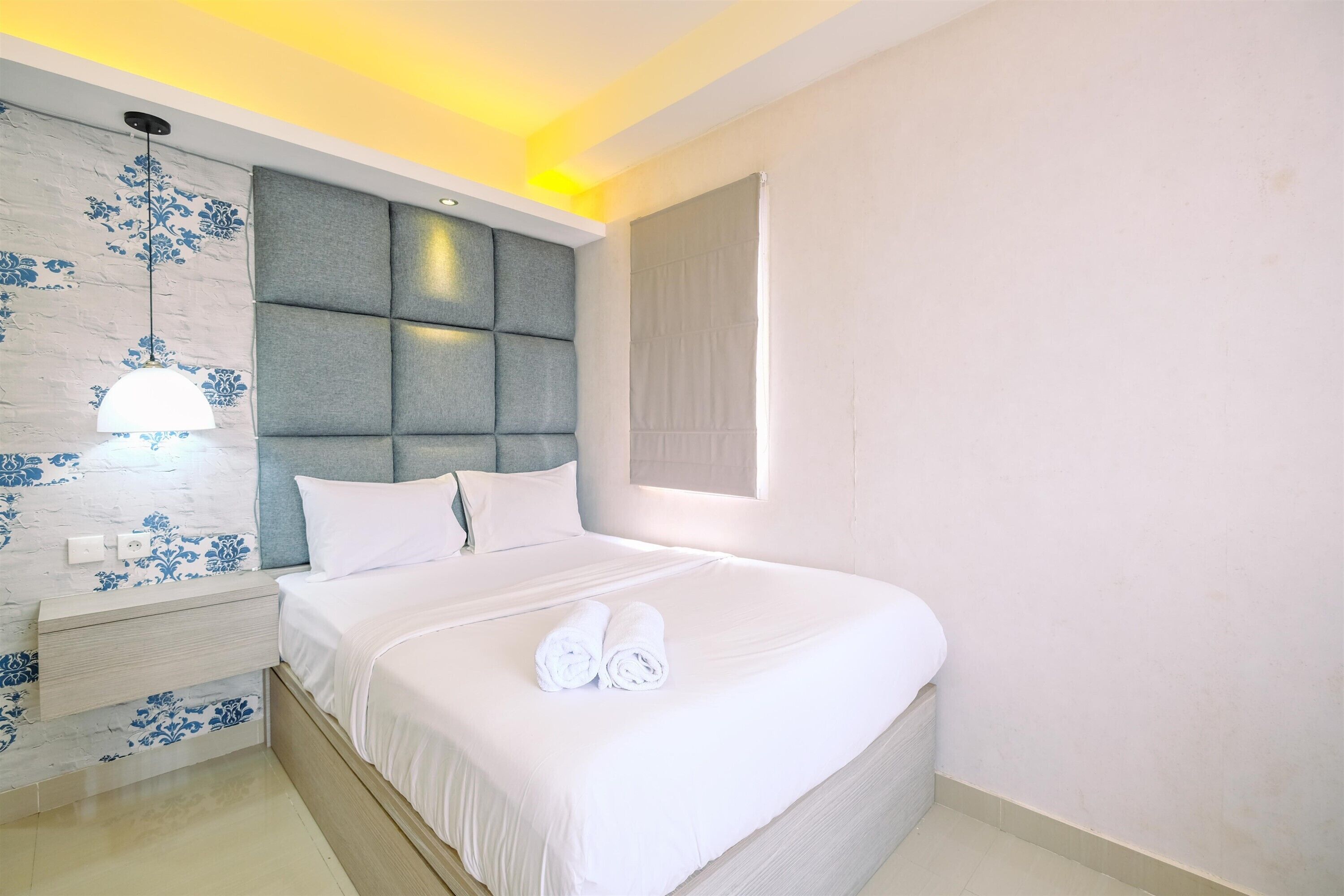 A neatly made bed with white linens and two rolled towels, a wall-mounted nightstand with a lamp, and a headboard with a padded design.