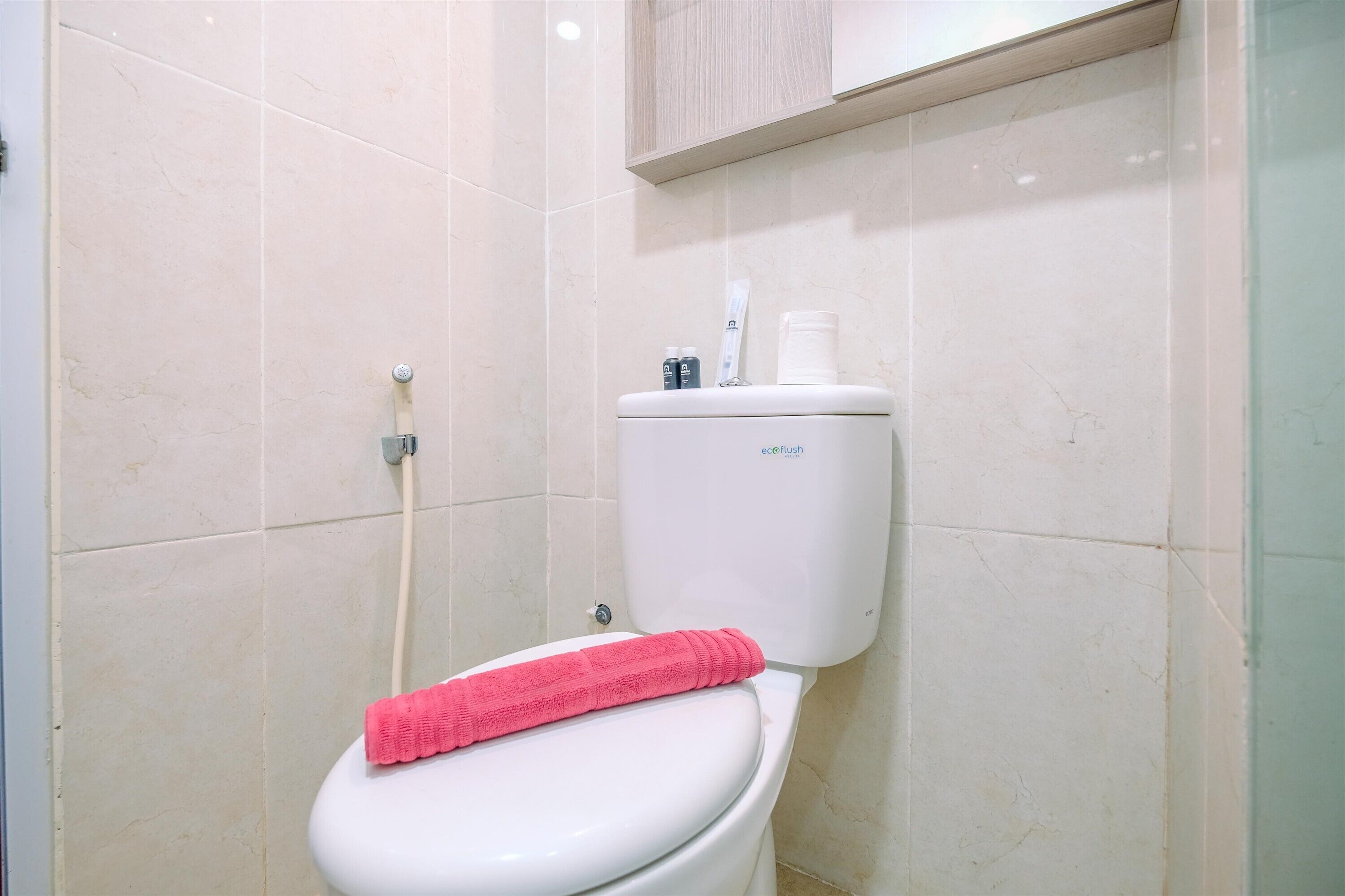 A bathroom with a white toilet, a pink towel on the seat, and a toilet paper holder with a roll and a bottle.