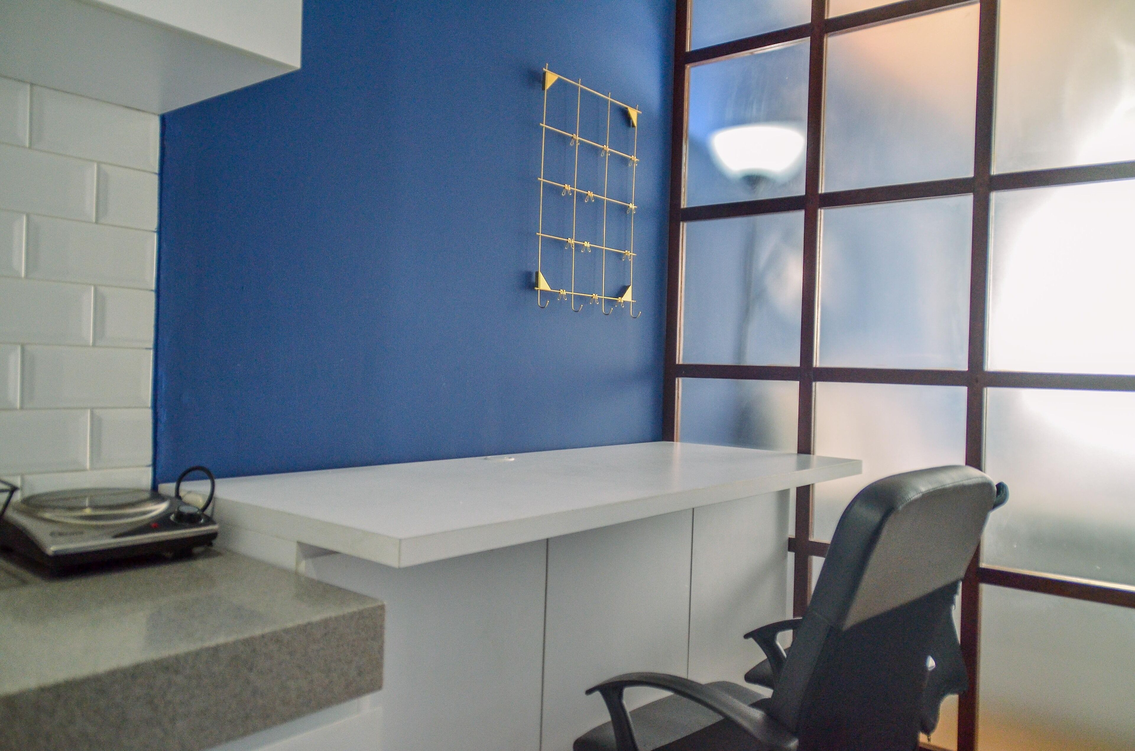 A room with a white desk, a blue accent wall, a window with blinds, and a chair.