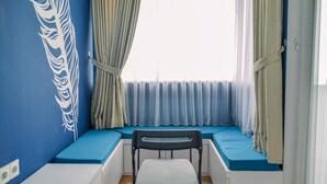 A room with a blue wall, a window with blue curtains, a white chair, and a bench with blue cushions. - Nice And Cozy Studio At Paddington Heights Apartment (Tangerang)
