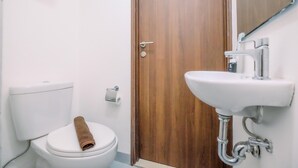A bathroom with a toilet, a wooden door, and a white sink with a mirror above it. - Modern Look Studio Lrt City Sentul - Royal Sentul Park Apartment (Bogor)