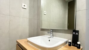 Un bagno moderno con un lavandino bianco, uno specchio e un mobile contenitore in legno. Sul mobile ci sono tre flaconi da bagno.