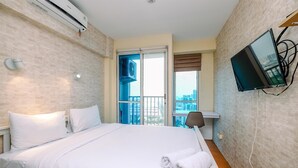 A hotel room with a bed, white linens, a TV mounted on the wall, and a view of the city through the window. - Minimalist Studio With Great City View At Tifolia Apartment (Jakarta)