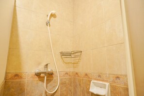 Área de duche com azulejos bege, chuveiro manual, torneira embutida na parede e um pequeno prateleira.