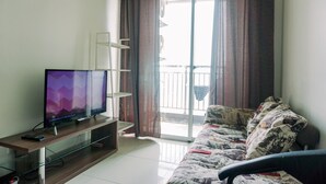 A living room with a flat-screen TV on a wooden stand, a sofa with patterned cushions, and a sliding glass door leading to a balcony.