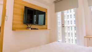 A hotel room with a mounted TV on a wooden panel, a large window with a view of buildings, and a door with a sliding glass panel. - Comfy Studio At 30Th Floor Tokyo Riverside Pik 2 Apartment (Teluknaga)