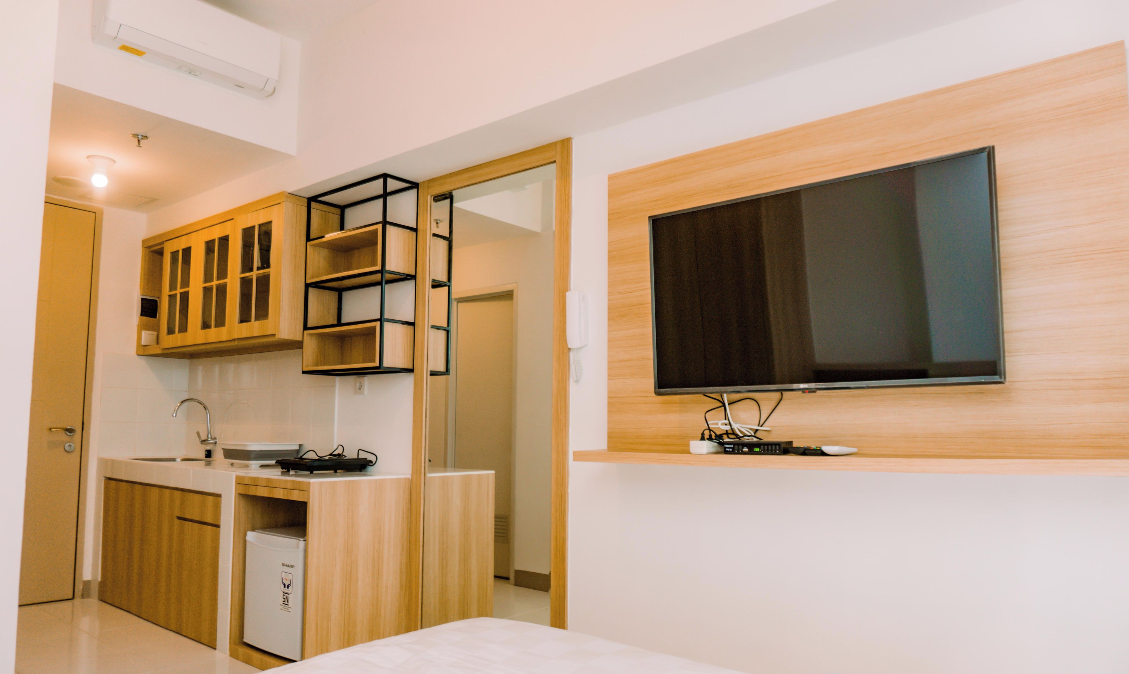 A modern hotel room with a kitchenette, a flat-screen TV mounted on the wall, and a bed.