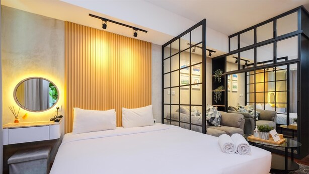 Une chambre d’hôtel moderne dotée d’un grand lit, d’une table de chevet avec une lampe, d’un miroir et d’une vue sur un espace salon séparé par une cloison en verre, comprenant un canapé et une table basse.