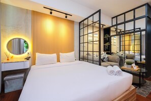 A modern hotel room with a large bed, a bedside table with a lamp, a mirror, and a view through a glass partition to a seating area with a sofa and a coffee table. - Simple And Comfort Studio Apartment At Nine Residence (Jakarta)