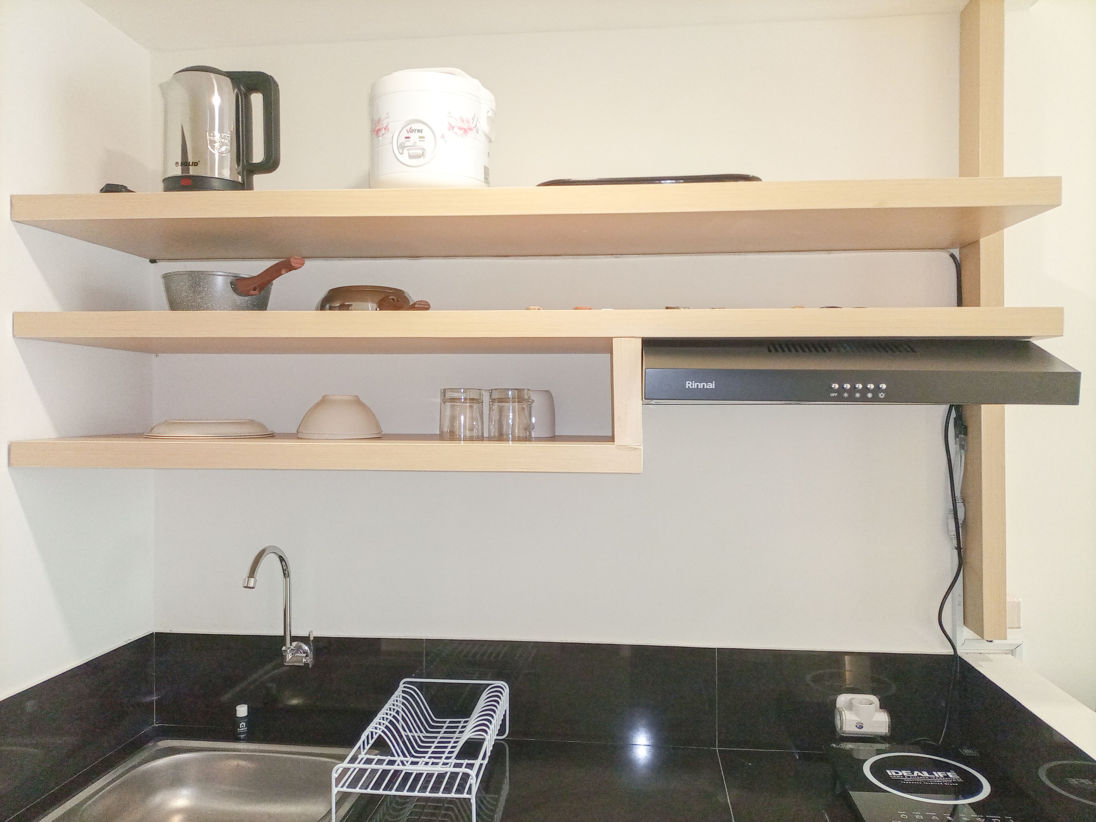 A kitchen with a black countertop, a sink, and a dish drying rack. There are wooden shelves above the countertop holding kitchen items.