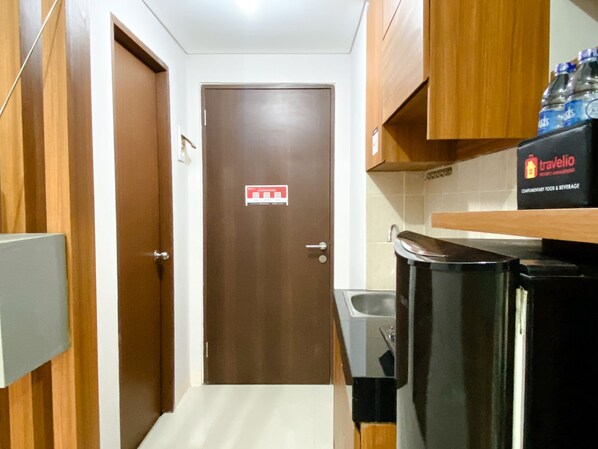 A narrow hallway with a brown door, a trash bin, a sink, and a shelf with bottled water.