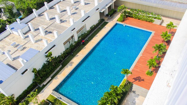 A rectangular swimming pool with a tiled deck, surrounded by greenery and a white building with a flat roof.