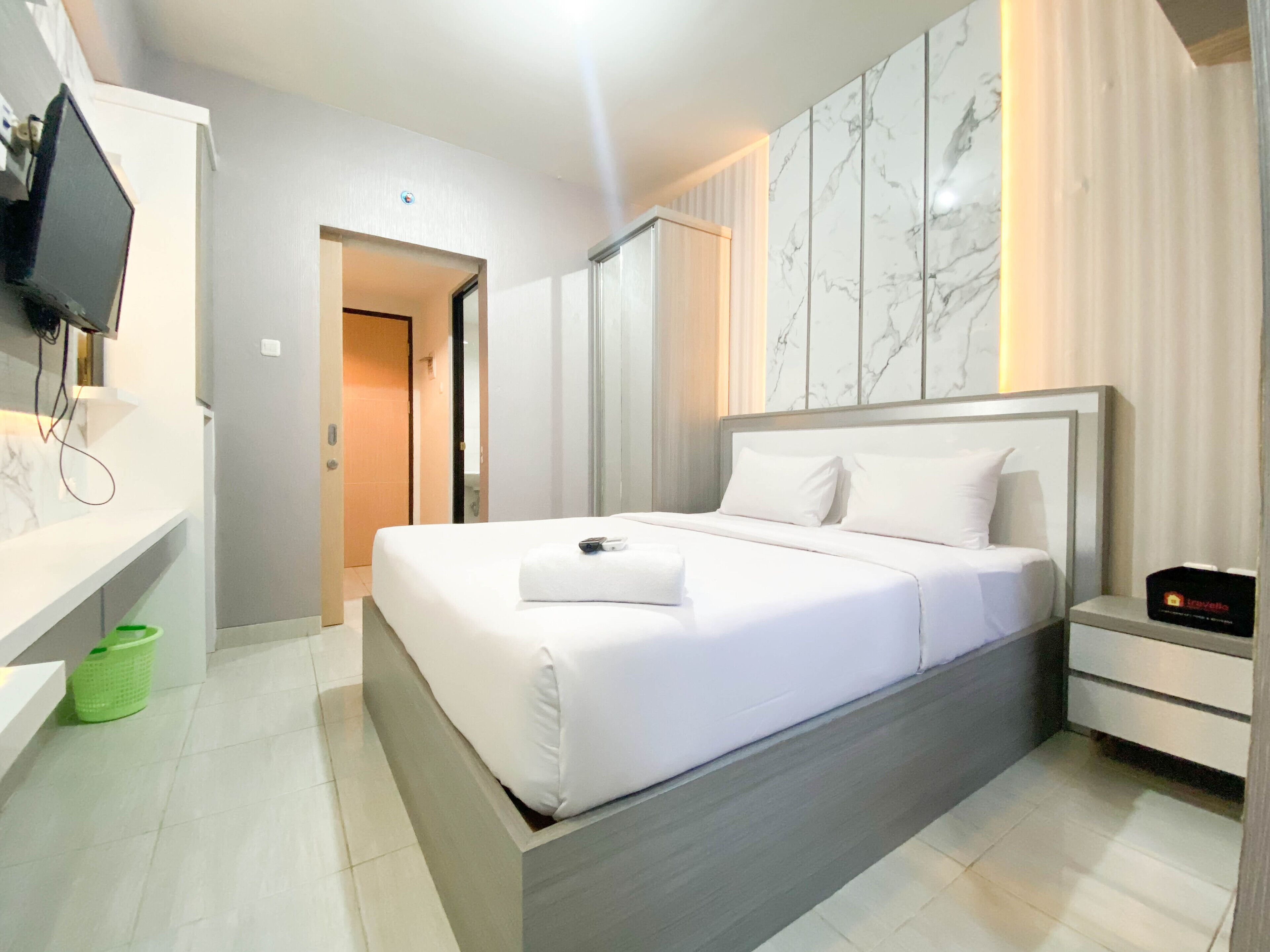 A modern hotel room with a large bed, a TV mounted on the wall, a bedside table, and a green laundry basket.
