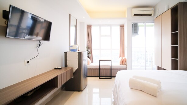 A modern hotel room with a bed, a TV mounted on the wall, a window with curtains, and an air conditioning unit. - Homey Studio At Taman Melati Surabaya Apartment (Surabaya)