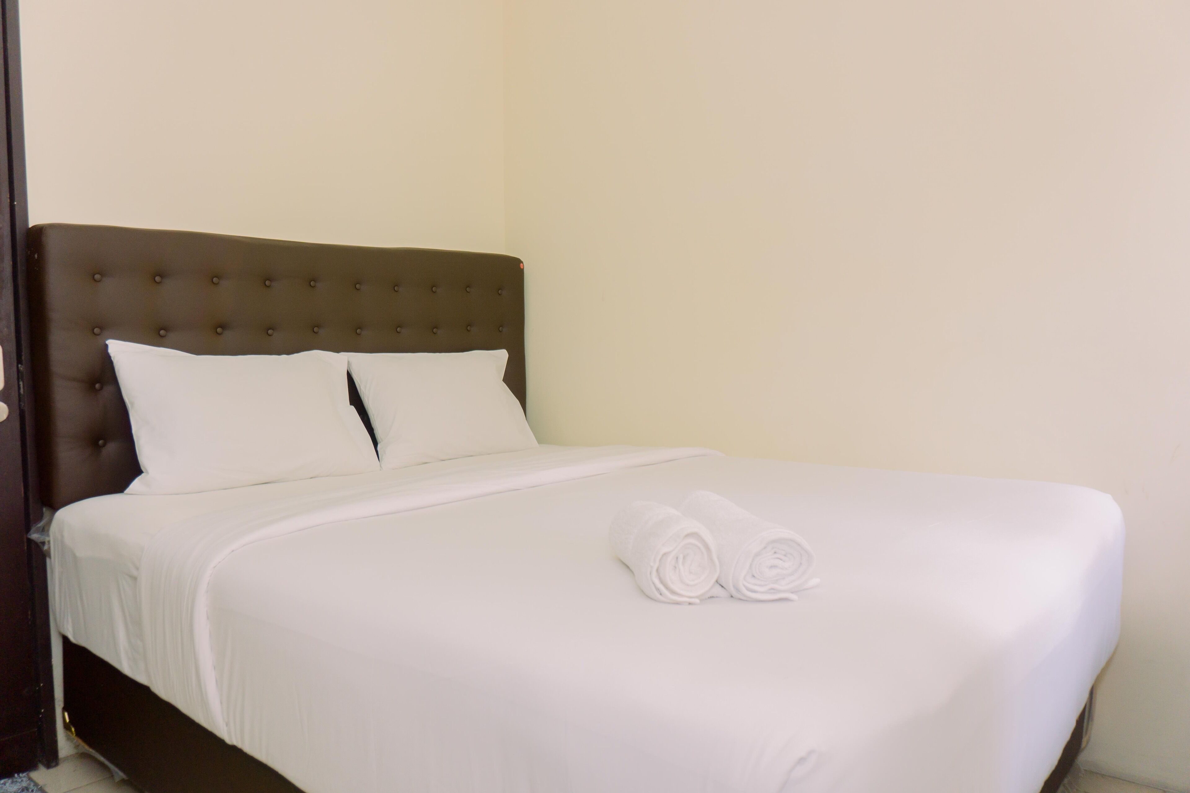 A neatly made bed with white linens and two rolled towels, a tufted headboard, and a plain wall in the background.