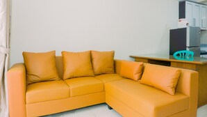 A modern living room with a large orange sectional sofa, a wooden coffee table, and a kitchen area with cabinets and appliances. - Modern Look And Homey 2Br At Great Western Resort Apartment (Tangerang)