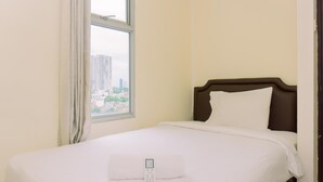 A neatly made bed with a remote control on it, a window with a view of buildings, and a plain wall. - Modern Look And Homey 2Br At Great Western Resort Apartment (Tangerang)