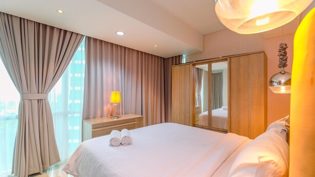 A hotel room with a large bed, a bedside table with a lamp, a mirror, and a view of the city. - Spacious 3Br At Kemang Village Apartment (Jakarta)