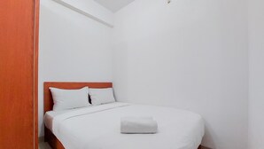 Una cama bien hecha con sábanas blancas y una toalla doblada, cabecera de madera y pared blanca.