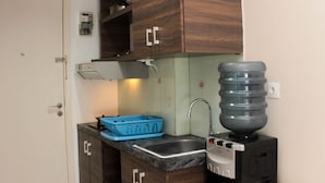 A compact kitchen with wooden cabinets, a sink, a water dispenser, and a blue dish rack. - Cozy Stay And Tranquil Studio Patraland Urbano Apartment (Bekasi)