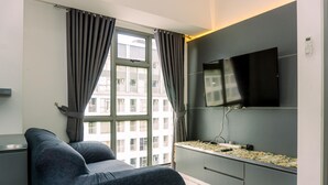 A modern living room with a grey sofa, a flat-screen TV mounted on the wall, and a window with curtains. - Comfort 1Br At 32Th Floor Apartment M-Town Residence (Gading Serpong)